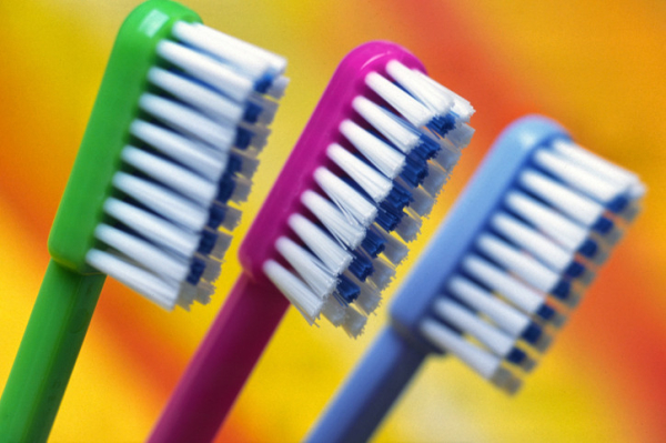 A trio of bright green, pink, and blue toothbrushes showing blue and white bristles in closeup against an orange and yellow background